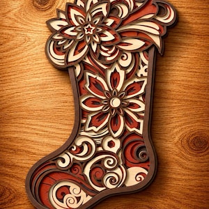 Op de afbeelding: Een meerlaagse kerstsokdecoratie van hout met ingewikkelde uitsparingen van bloemmotieven en krullend vlechtwerk. Het ontwerp maakt gebruik van een kleurenpalet van donkerbruin, warm rood en crème, wat een ruimtelijk effect creëert.