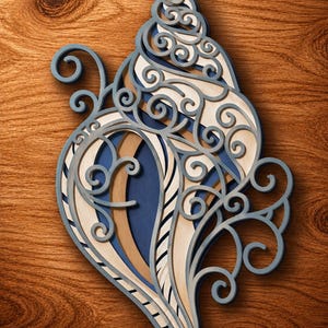 May include: A multi-layered wooden wall art piece shaped like a seashell, featuring intricate laser-cut scrollwork patterns in shades of navy blue, light gray, and natural wood tones, creating a dimensional effect with stacked cutouts.
