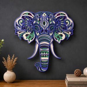 May include: A multi-layered wooden wall art piece shaped like an elephant head. The design features intricate cutouts in shades of deep blue, forest green, and cream, creating a dimensional effect with ornate patterns across the ears and trunk.