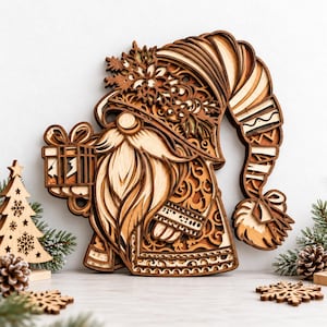 May include: A multi-layered wooden wall art piece depicting a gnome with a long beard, wearing a tall, patterned hat adorned with snowflake and holly motifs, holding a gift box, with a separate wooden Christmas tree ornament nearby.