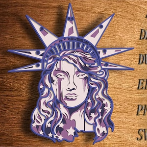 May include: A purple and white layered paper cut design of the Statue of Liberty. The design features a detailed face and crown with stars.