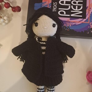 Könnte beinhalten: Eine gehäkelte Puppe mit schwarzen Haaren, die einen schwarzen Mantel, weiß-schwarz gestreifte Socken und schwarze Schuhe mit weißen Verzierungen trägt. Die Puppe steht auf einer weißen Oberfläche. Die Puppe hält eine kleine schwarze Tasche.