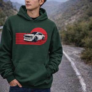 Op de afbeelding: Donkergroene hoodie met een afbeelding van een witte auto voor een rood zonontwerp. De hoodie heeft een voorzak en een capuchon. De persoon draagt een blauwe spijkerbroek en staat op een weg.