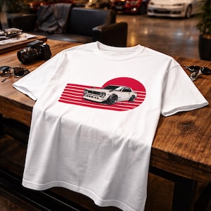 May include: White t-shirt featuring a graphic of a classic white car with black wheels, set against a red circle and horizontal red stripes. The design evokes a vintage automotive theme, perfect for car enthusiasts.