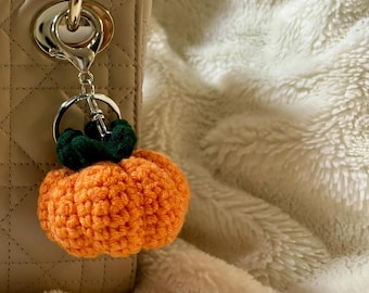 May include: A crochet pumpkin keychain with a bright orange body and dark green stem, attached to a silver-toned metal lobster clasp and ring, hanging from the side of a beige quilted handbag with a structured handle and silver hardware.