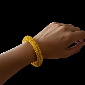 May include: A bright yellow beaded bracelet. The bracelet is worn on a person's wrist against a black background. The bracelet is made of small, round beads, and the color is a vibrant, sunny yellow.