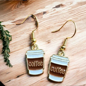 Peut inclure: Boucles d'oreilles pendantes dorées avec un charme en forme de tasse à café blanche. Le charme a le mot "café" imprimé en brun sur le côté.