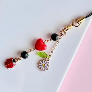 May include: A white phone case with a black and gold phone charm. The charm features a red heart, a green leaf, a white daisy, and a red ladybug.