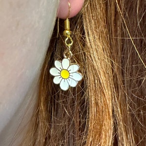 Small, dainty white daisy flower earrings with yellow centers, dangling from gold earring hooks. Displayed in an earlobe to show size.