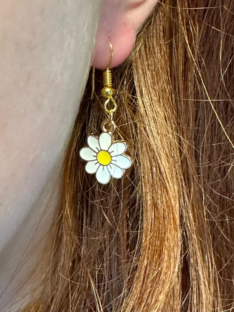 Small, dainty white daisy flower earrings with yellow centers, dangling from gold earring hooks. Displayed in an earlobe to show size.