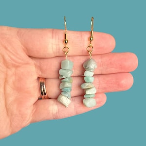 May include: A pair of gold dangle earrings with light green gemstone chips. The earrings are made with a gold hook and a chain of small, irregular shaped green stones.