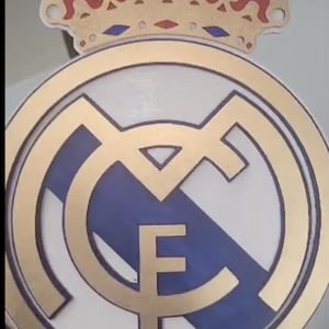 May include: A gold and white circular emblem with a blue and white crest featuring a stylized "M" and a gold "F". The emblem is topped with a gold crown with red and white accents.