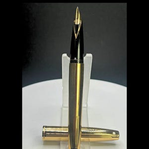 May include: A fountain pen featuring a gold-toned metal body with vertical pinstripes and a black section leading to a gold nib. The pen is displayed upright with its matching gold-toned metal cap resting horizontally at the base of the stand.