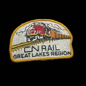 May include: Embroidered patch depicting a train with a red engine and orange carriages on a yellow track. The patch reads "CN RAIL GREAT LAKES REGION" in black text on a white background, with a yellow border.