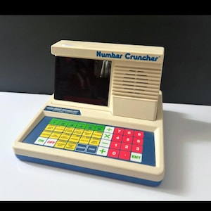 May include: Vintage "Number Cruncher" toy calculator in off-white and blue. Features a keyboard with colored keys, a dark screen, and the text "Number Cruncher". A retro educational toy.