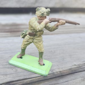 May include: A vintage toy soldier figurine, painted in shades of khaki and olive green, aiming a rifle. The soldier wears a helmet and uniform, standing on a green rectangular base. The toy has a weathered appearance, suggesting it is an antique.