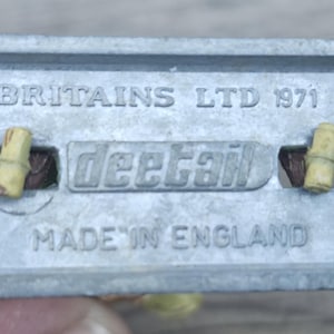 May include: A close-up of the underside of a vintage toy component. The metal piece is stamped with "BRITAINS LTD 1971", "deetail", and "MADE IN ENGLAND". Two small, cylindrical pieces are visible.