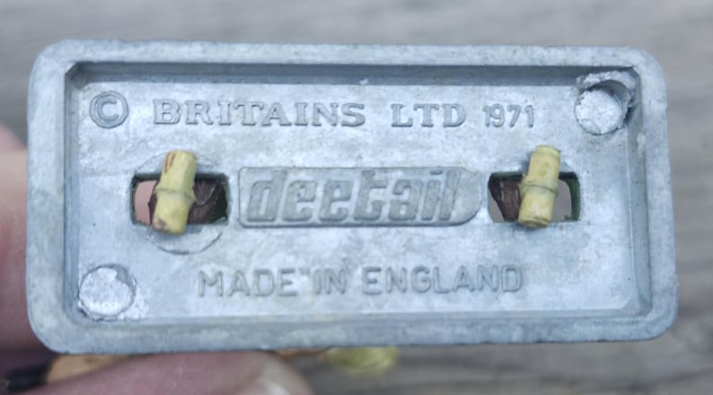 May include: A close-up of the underside of a vintage toy component. The metal piece is stamped with "BRITAINS LTD 1971", "deetail", and "MADE IN ENGLAND". Two small, cylindrical pieces are visible.