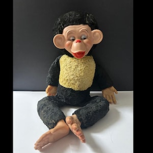 May include: A vintage monkey doll with a black furry body, arms, and legs. The doll has a yellow chest, a painted face with large ears, and a red mouth. The doll is sitting with its legs crossed and its hands resting on its legs.