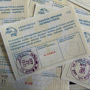 May include: A pile of vintage international reply coupons. The coupons are cream-coloured with blue and red text and stamps. The stamps indicate the origin and date, such as Honolulu, September 17, 1977.