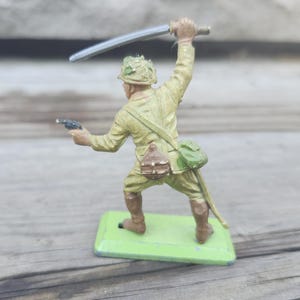 May include: A vintage toy soldier figurine in a combat pose, wielding a sword and pistol. The figure is painted in olive drab with brown boots and a green base. The soldier has a helmet and a satchel.