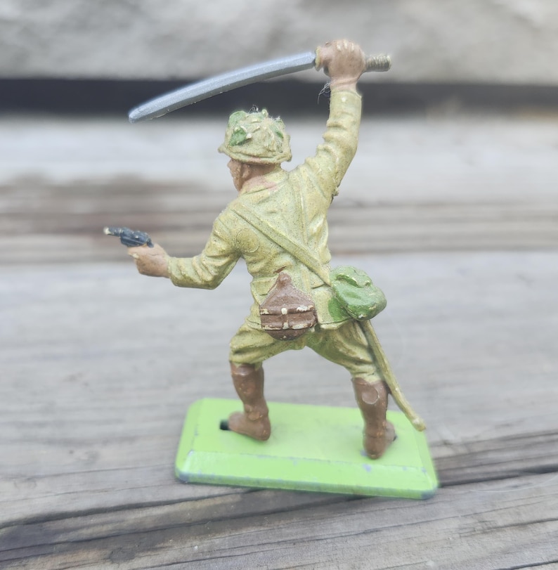 May include: A vintage toy soldier figurine in a combat pose, wielding a sword and pistol. The figure is painted in olive drab with brown boots and a green base. The soldier has a helmet and a satchel.