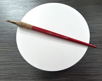 May include: A red dip pen with a cork grip and a metallic nib. The barrel features the white printed text 77 E. FABER U.S.A. ECHO. The pen is positioned diagonally across a circular white surface, showcasing its slender design and classic construction.