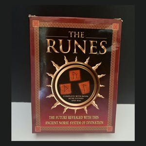 May include: A boxed set titled "THE RUNES" with a dark red and gold design. The box features a central circular emblem with rune stones and text that reads "THE FUTURE REVEALED WITH THIS ANCIENT NORSE SYSTEM OF DIVINATION."