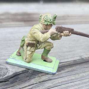 May include: A vintage toy soldier figurine in a kneeling position, holding a rifle. The soldier is painted in olive green and khaki tones, with a green helmet and a green base. The toy is likely made of metal.