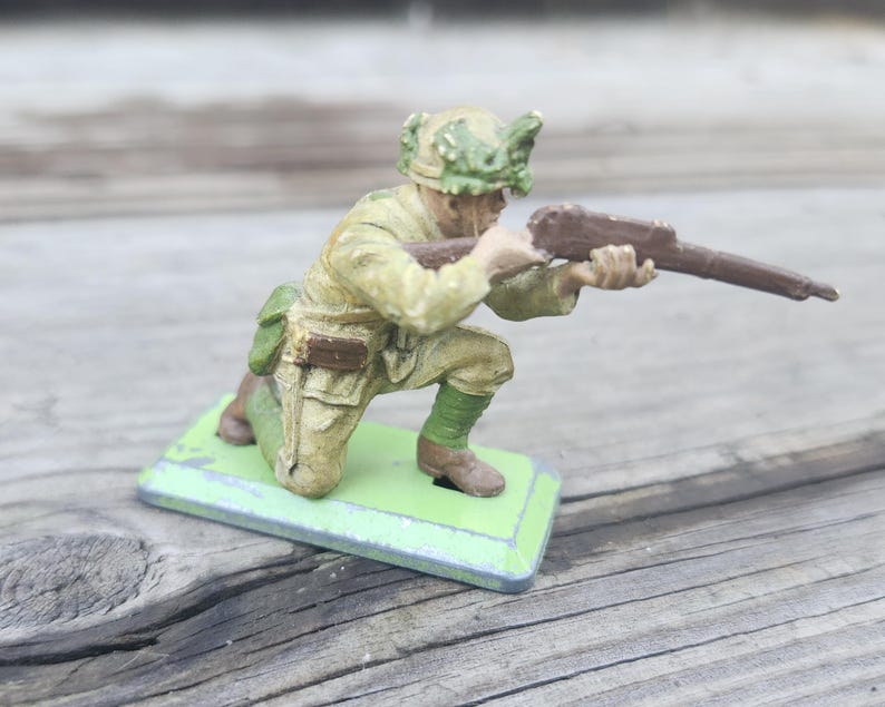 May include: A vintage toy soldier figurine in a kneeling position, holding a rifle. The soldier is painted in olive green and khaki tones, with a green helmet and a green base. The toy is likely made of metal.