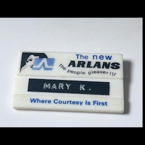 May include: A rectangular white name tag with blue text and a graphic. The top section reads "The new ARLANS the people pleaser!!" with a logo. The middle section says "MARY K." and the bottom says "Where Courtesy Is First."