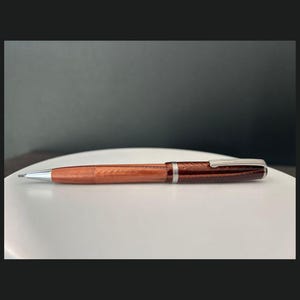 May include: A wooden pen with a silver clip and tip. The pen has a light brown barrel and a dark brown, textured cap. The pen is lying on a white surface against a dark gray background.