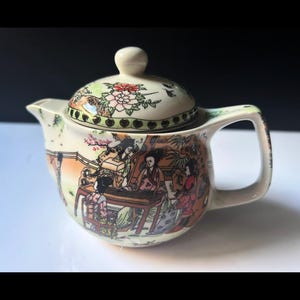 May include: A small, cream-coloured ceramic teapot with a lid. The lid is decorated with floral patterns. The body of the teapot features a detailed illustration of people in a traditional setting. The handle and spout are curved.
