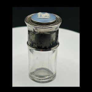 May include: A small, antique glass jar with a silver-colored metal top. The top features a light blue circular design with a white relief image. The jar is cylindrical and appears to be a container for a small amount of liquid or powder.