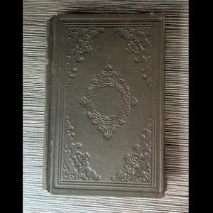 May include: A vintage, dark green book with ornate embossed floral and geometric designs. The cover features a central diamond-shaped frame and decorative corner elements. The book's edges show signs of wear, suggesting its age.