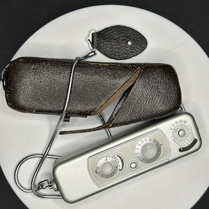 May include: A vintage silver light meter with dials and a leather case. The meter has a silver chain and a small leather piece attached. The case is brown and textured.