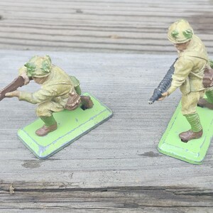 May include: Two vintage toy soldiers in olive drab uniforms, one kneeling and aiming a rifle, the other standing with a machine gun. Both figures are on green rectangular bases, set against a weathered wooden surface.