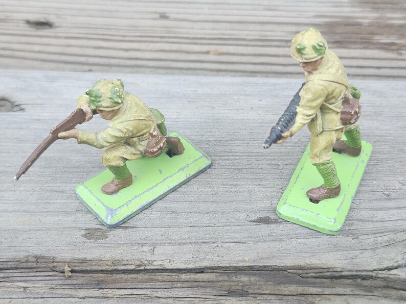 May include: Two vintage toy soldiers in olive drab uniforms, one kneeling and aiming a rifle, the other standing with a machine gun. Both figures are on green rectangular bases, set against a weathered wooden surface.
