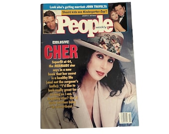 May include: A People Weekly magazine cover featuring Cher. The cover includes the headline "EXCLUSIVE! CHER" and text about her new book. Cher is wearing a hat with floral accents and a light-coloured blazer.
