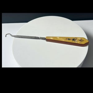 May include: An antique dental tool with a curved metal end and a decorative handle. The handle is a combination of brown and yellow, with small black and gold embellishments. The tool is placed on a white surface.