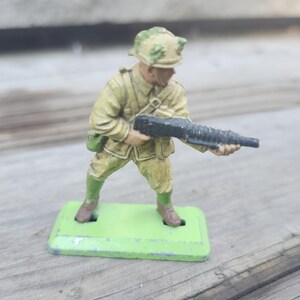 May include: A vintage toy soldier figurine in a khaki uniform and helmet, holding a black rifle. The soldier stands on a green rectangular base. The figurine is painted with details and has a weathered appearance.