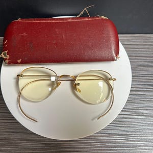 May include: Vintage gold-tone eyeglasses with oval lenses and wire temples, resting on a white surface. A worn, red leather eyeglasses case sits above the glasses. The glasses have a classic, retro design.