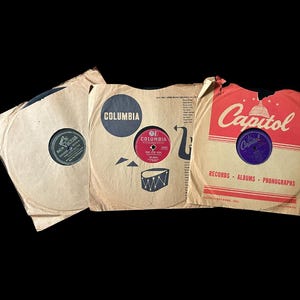 May include: Three vintage vinyl record albums in worn paper sleeves. The sleeves feature the logos "Columbia" and "Capitol Records". The records are in various colors, including red and purple, and are set against a black background.