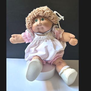 May include: A vintage Cabbage Patch Kids doll with blonde yarn hair and green eyes. The doll is dressed in a pink and white dress, matching bloomers, and white shoes. The doll is seated on a white pedestal.