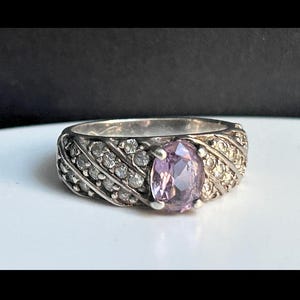 May include: Silver ring with a large oval, light purple gemstone. The band is detailed with small, clear stones and a textured design. The ring is set against a white surface with a dark background.