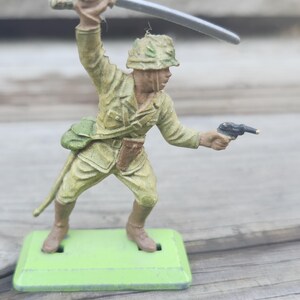 May include: A vintage toy soldier figurine in a combat pose, wielding a sword and pistol. The figure is painted in olive green and brown tones, with a green base. The soldier is wearing a helmet and military uniform.
