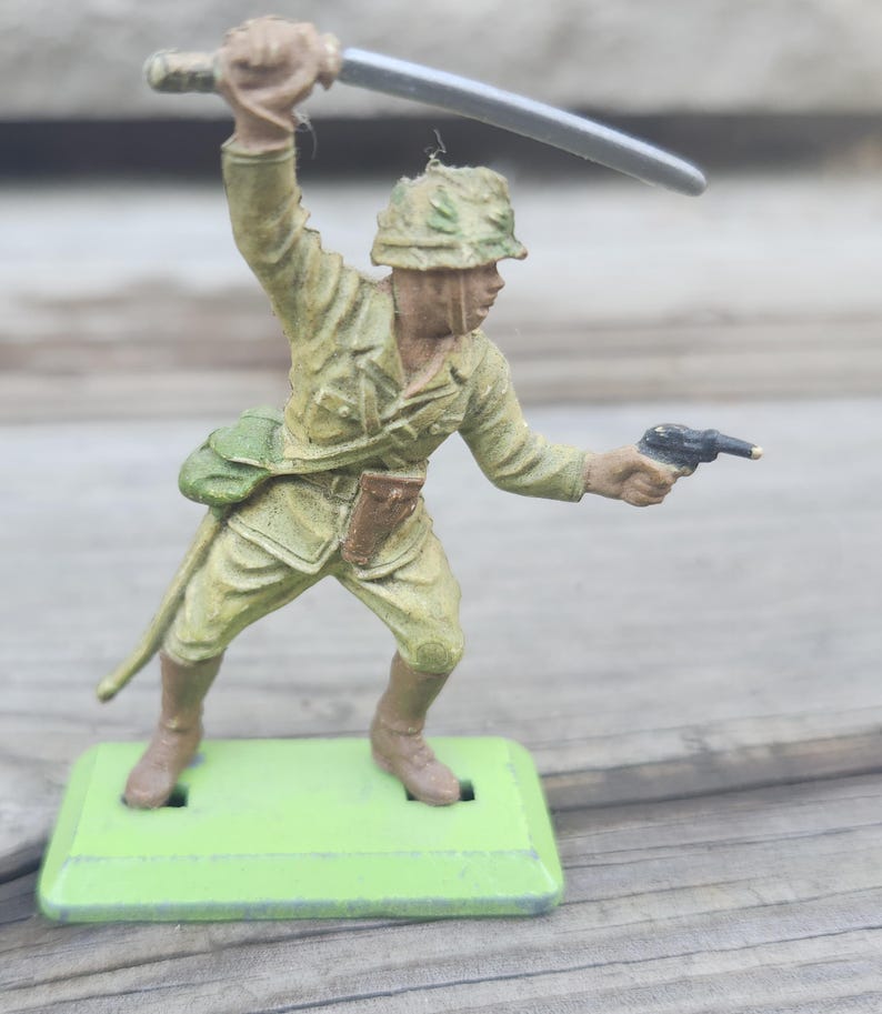 May include: A vintage toy soldier figurine in a combat pose, wielding a sword and pistol. The figure is painted in olive green and brown tones, with a green base. The soldier is wearing a helmet and military uniform.