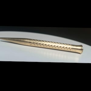 Peut inclure: Un porte-mine en métal doré doté d'une pointe effilée et d'un corps cylindrique orné d'un motif répétitif en chevrons ou en arêtes de poisson. L'extrémité du corps présente une section de capuchon lisse et légèrement évasée.