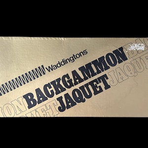 May include: A gold-coloured box featuring the text "Waddingtons BACKGAMMON JAQUET" in black and white. The box has a diagonal striped design and is set against a black background. The product is a board game.