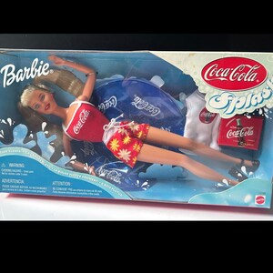 May include: A Barbie doll in a Coca-Cola Splash themed box. The doll is dressed in a red top with the Coca-Cola logo and floral shorts. The box features the Coca-Cola logo and a blue water splash design. Includes a white t-shirt with the Coca-Cola logo.